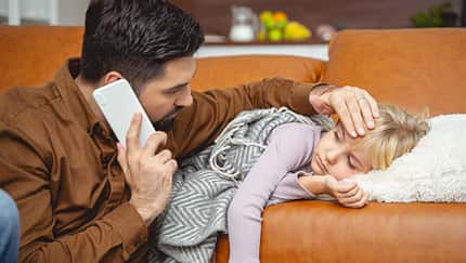 Ein Vater kümmert sich um seine kranke Tochter, die auf dem Sofa liegt und telefoniert gleichzeitig