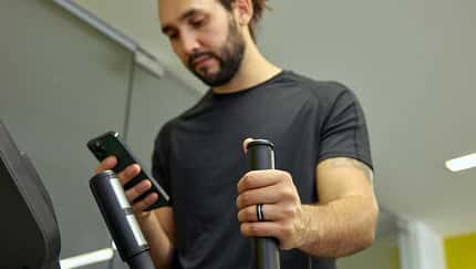 Ein Mann trainiert auf einem Crosstrainer im Fitnessstudio, hält einen Griff des Geräts und blickt auf sein Smartphone. Er trägt einen Fitnessring