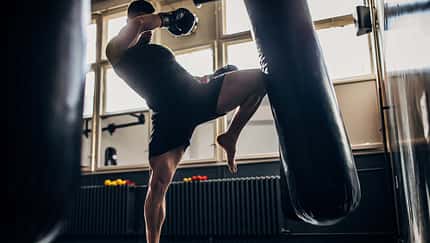 Mann trainiert Kampfsport im Boxstudio und tritt mit einem Bein gegen einen Boxsack.