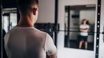 Mann steht mit dem Rücken zum Betrachter in einem leeren Fitnessstudio vor dem Spiegel