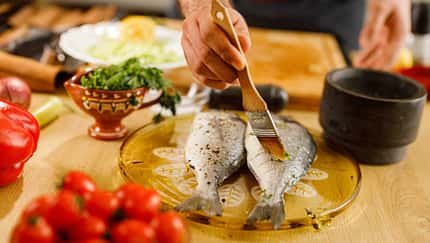 Person bestreicht zwei rohe Fische mit Marinade auf einem Küchentisch mit frischem Gemüse. Person bestreicht zwei rohe Fische mit Marinade auf einem Küchentisch mit frischem Gemüse.