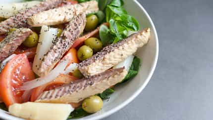 Schüssel mit Salat aus Tomaten, grünen Oliven, Zwiebeln und Blattgrün, belegt mit Sardinenfilets und bestreut mit Chiasamen.
