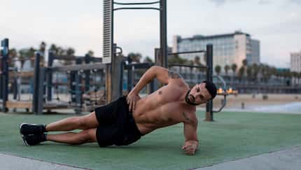Mann in Sportshorts macht seitliche Plank-Übung auf einer Outdoor-Trainingsfläche am Strand, im Hintergrund Fitnessgeräte und Gebäude. Mann in Sportshorts macht seitliche Plank-Übung auf einer Outdoor-Trainingsfläche am Strand, im Hintergrund Fitnessgeräte und Gebäude.