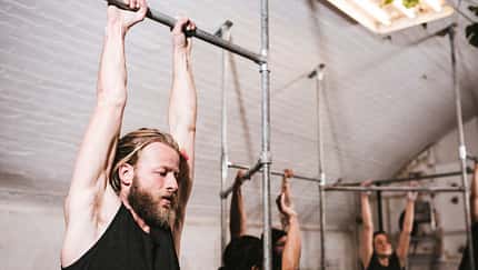 Mann hängt mit gestreckten Armen an einer Klimmzugstange in einem Fitnessstudio, im Hintergrund trainieren weitere Personen.