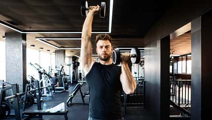 Mann im Fitnessstudio führt mit Kurzhanteln eine Schulterübung aus, ein Arm über Kopf gestreckt, der andere auf Schulterhöhe. Mann im Fitnessstudio führt mit Kurzhanteln eine Schulterübung aus, ein Arm über Kopf gestreckt, der andere auf Schulterhöhe.