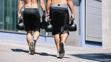 Zwei Männer gehen draußen auf der Straße und tragen jeweils schwere Kettlebells in beiden Händen.