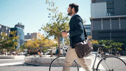 Geschäftsmann schiebt mit Kaffeebecher und Umhängetasche sein Fahrrad durch die Stadt