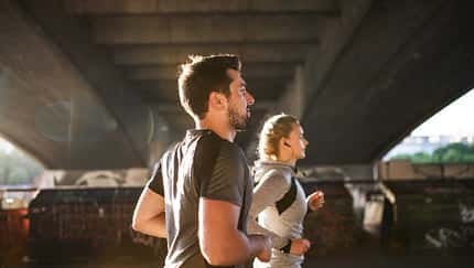 Mann und Frau joggen unter einer Brücke durch, die Sonne im Rücken