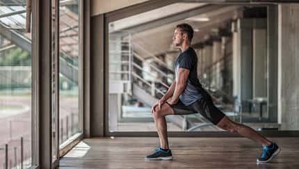 Sportlicher Mann macht einen Ausfallschritt-Stretch in einer modernen Trainingshalle vor großen Fenstern.