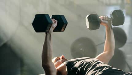 Mann liegt auf einer Trainingsbank und führt im Fitnessstudio eine Kurzhantel-Bankdrück-Übung aus.