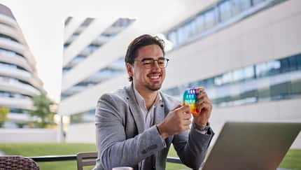 Mann sitzt mit Laptop im Freien und spielt lächelnd mit einem bunten Pop-It-Fidget-Toy.