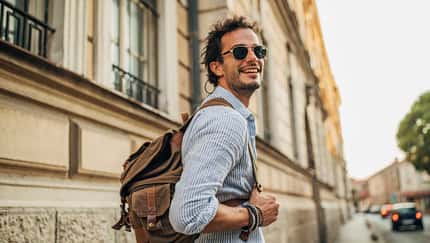 Junger Mann mit Sonnenbrille und Rucksack spaziert und dreht sich lächelnd um.