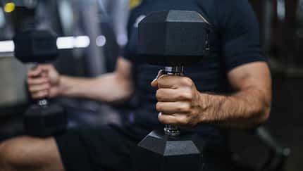 Eine Person sitzt auf einer Bench und hält zwei Hanteln in den Händen. Fokus auf eine Hand mit der Hantel