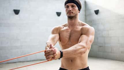 Ein Mann trainiert oberkörperfrei in einem Raum und hält mit beiden Armen ein Stretching-Band