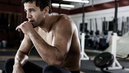 Sportlicher Mann in Sportleggings und oberkörperfrei sitzt im Fitnessstudio in der Hocke und sieht erschöpft aus