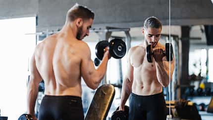 Junger muskulöser Mann macht Kurzhantel-Übungen im Fitnessstudio vor einem Spiegel