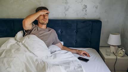 Junger Mann sitzt im Bett und hält sich seinen Kopf