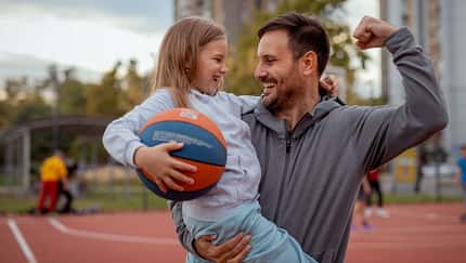 Ein Mann, der einen Ball und seine Tochter im Arm hält und seinen Bizeps anspannt