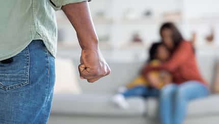 Im Vordergrund sieht man eine geballte Männerfaust, im Hintergrund eine verängstigte Mutter und ihr Kind, die auf einem Sofa sitzen.