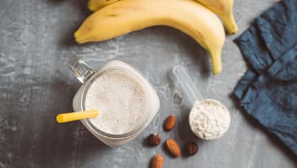 Proteinsmoothie im Glas mit Mandeln, Banane und einem Scoop Proteinpulver daneben