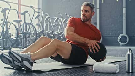 Mann führt eine Bauch-Übung mit einem Ball auf einet Sportmatte aus, im Hintergrund sind Sportgeräte zu sehen