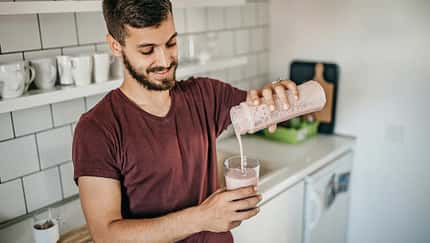 Junger Mann steht in der Küche und schenkt sich einen frisch gemixten Proteinshake in ein Glas ein