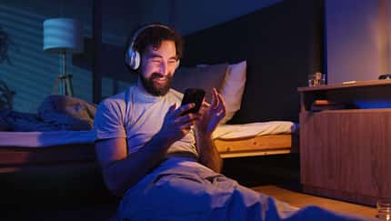 Mann mit Kopfhörern sitzt im dunklen Schlafzimmer an sein Bett gelehnt und schaut lachend auf sein Smartphone Mann mit Kopfhörern sitzt im dunklen Schlafzimmer an sein Bett gelehnt und schaut lachend auf sein Smartphone