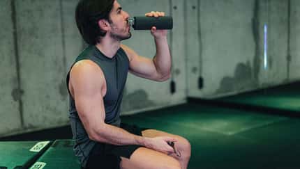 Mann sitzt im Fitnessstudio auf einer Fitnessbank und trinkt aus seiner Trinkflasche Mann sitzt im Fitnessstudio auf einer Fitnessbank und trinkt aus seiner Trinkflasche
