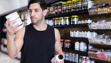 Ein Mann steht vor einem Regal mit Protein-Supplementen und schaut sich die Inhaltsstoffe von zwei Produkten an Ein Mann steht vor einem Regal mit Protein-Supplementen und schaut sich die Inhaltsstoffe von zwei Produkten an