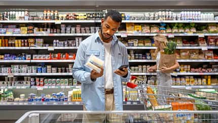 Mann steht im Supermarkt vor dem Kühlregal und schaut prüfend auf die Rückseite eines Produktes Mann steht im Supermarkt vor dem Kühlregal und schaut prüfend auf die Rückseite eines Produktes