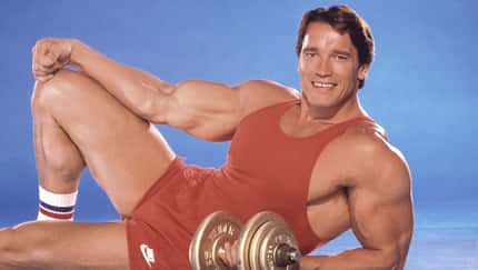 Arnold Schwarzenegger posiert vor einem blauen Hintergrund in einem roten Sportoutfit mit einer Kurzhantel Arnold Schwarzenegger posiert vor einem blauen Hintergrund in einem roten Sportoutfit mit einer Kurzhantel