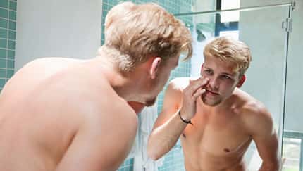 Ein Mann schaut sich oberkörperfrei im Spiegel seine Augenringe an
