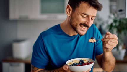 Ein Mann im blauen Sportshirt isst glücklich Haferflocken mit Obst aus einer Schale