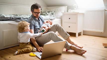 Ein Vater sitzt vor einem Sofa, mit einem aufgeklappten Notebook und zwei kleinen Kindern an seiner Seite Ein Vater sitzt vor einem Sofa, mit einem aufgeklappten Notebook und zwei kleinen Kindern an seiner Seite