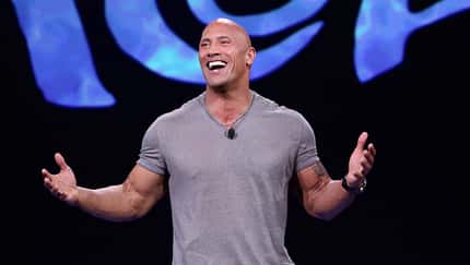 Dwayne Johnson im blaugrauen Shirt mit ausgebreiteten Armen, der auf der Premiere von "Moana" in die Kamera lacht Dwayne Johnson im blaugrauen Shirt mit ausgebreiteten Armen, der auf der Premiere von "Moana" in die Kamera lacht