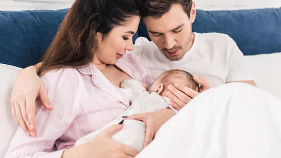 Eine frischgebackene Mutter sitzt aufrecht im Bett und gibt ihrem Baby die Brust. Der Vater sitzt daneben und schaut zu