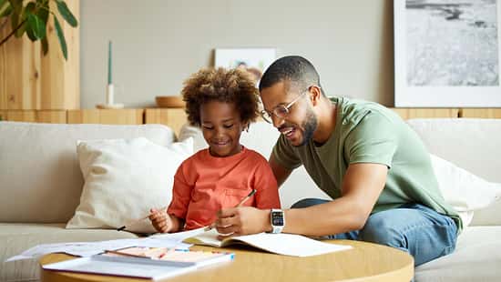 Ein Vater sitzt mit seiner Tochter am Wohnzimmertisch und hilft ihr bei den Hausaufgaben