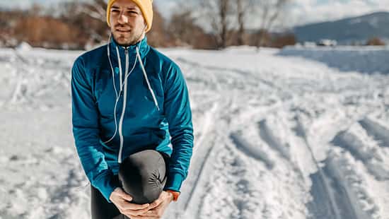 Mann in Laufkleidung steht in verschneitem Park und dehnt sich