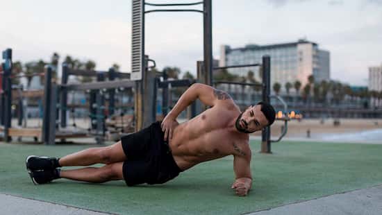 Mann in Sportshorts macht seitliche Plank-Übung auf einer Outdoor-Trainingsfläche am Strand, im Hintergrund Fitnessgeräte und Gebäude.