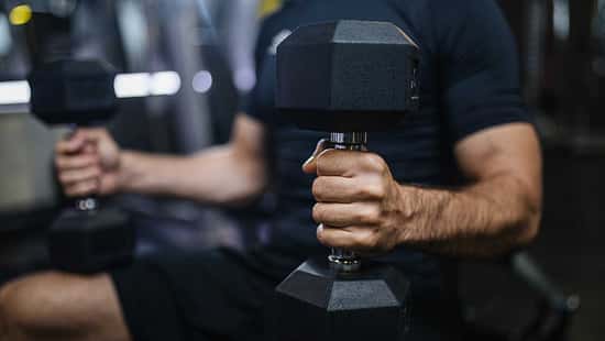 Eine Person sitzt auf einer Bench und hält zwei Hanteln in den Händen. Fokus auf eine Hand mit der Hantel
