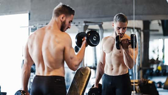 Junger muskulöser Mann macht Kurzhantel-Übungen im Fitnessstudio vor einem Spiegel