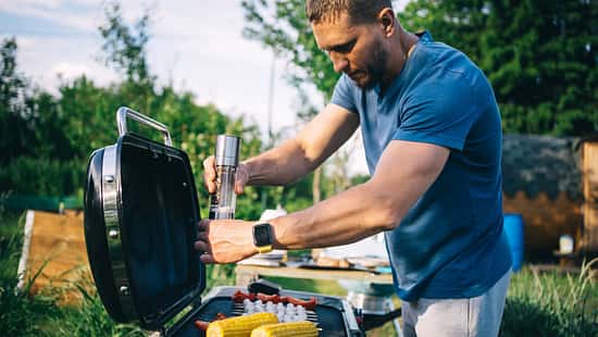 Ein Mann steht am Grill und würzt Maiskolben