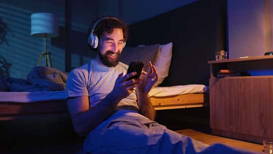 Mann mit Kopfhörern sitzt im dunklen Schlafzimmer an sein Bett gelehnt und schaut lachend auf sein Smartphone