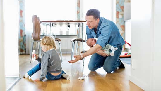 Ein junger Vater mit Säugling auf dem Arm hockt vor seiner kleinen Tochter auf dem Boden