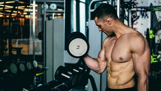Ein trainierter Mann steht oberkörperfrei im Gym und beobachtet seinen großen Bizeps, während er ihn mit Kurzhanteln trainiert