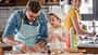 Ein Vater und sein Kind backen in der Küche, im Hintergrund sieht man die Mutter