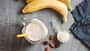 Proteinsmoothie im Glas mit Mandeln, Banane und einem Scoop Proteinpulver daneben