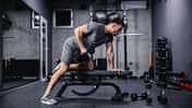 Ein muskulöser Mann trainiert mit einer Hantel, die sich auf einer Sportbank im Fitnessraum befindet. 