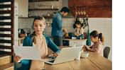 Eine Mutter sitzt am Esstisch in der Küche und arbeitet am Notebook, im Hintergrund sieht man die zwei Töchter und den Mann, der gerade das Essen zubereitet