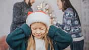 Ein Mädchen mit Weihnachtsmütze hält sich die Ohren zu, im Hintergrund sieht man, wie sich die Eltern lautstark streiten